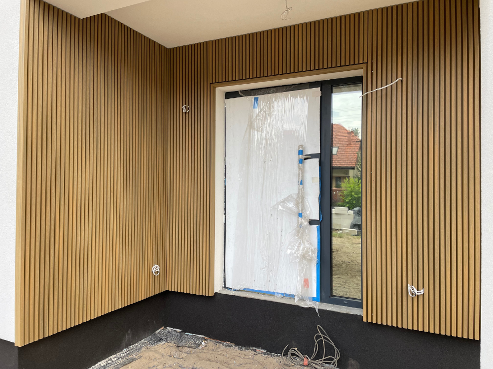 Lamele elewacyjne UltraShield® Naturale w kolorze Oak zamontowane na ścianie w podcieniu przy drzwiach wejściowych do budynku jednorodzinnego